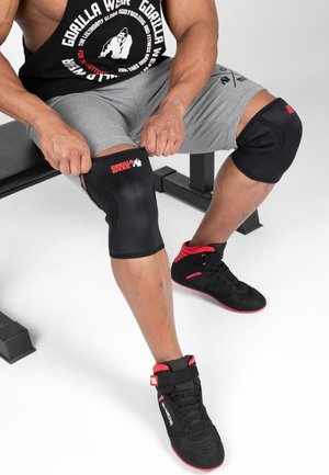 Homme portant des baskets noires et rouges, un short gris et un débardeur noir avec des genouillères assis sur un banc.