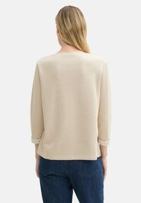 Beige Oberteil mit geripptem Muster, Dreiviertelärmeln, runder Ausschnitt und hochgekrempelten Bündchen, kombiniert mit dunklen Jeans. Strukturiertes Gewebedetail sichtbar.