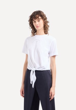 T-shirt en coton blanc avec un détail noué à l'avant. Manches courtes et décolleté rond, avec un imprimé graphique subtil sur la zone de la poitrine.