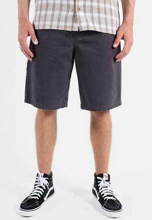 Homme portant un short sombre jusqu'aux genoux, des baskets montantes noires avec des lacets blancs, et une chemise à carreaux beige.