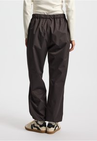 Pantaloni impermeabili marroni con vita elasticizzata, caratterizzati da una vestibilità ampia e una texture liscia, abbinati a sneakers di colore chiaro.
