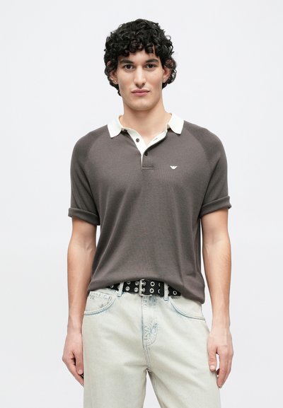 Polo gris avec un col blanc et deux boutons, manches courtes retroussées et petit logo sur le côté gauche de la poitrine, porté avec un jean clair.