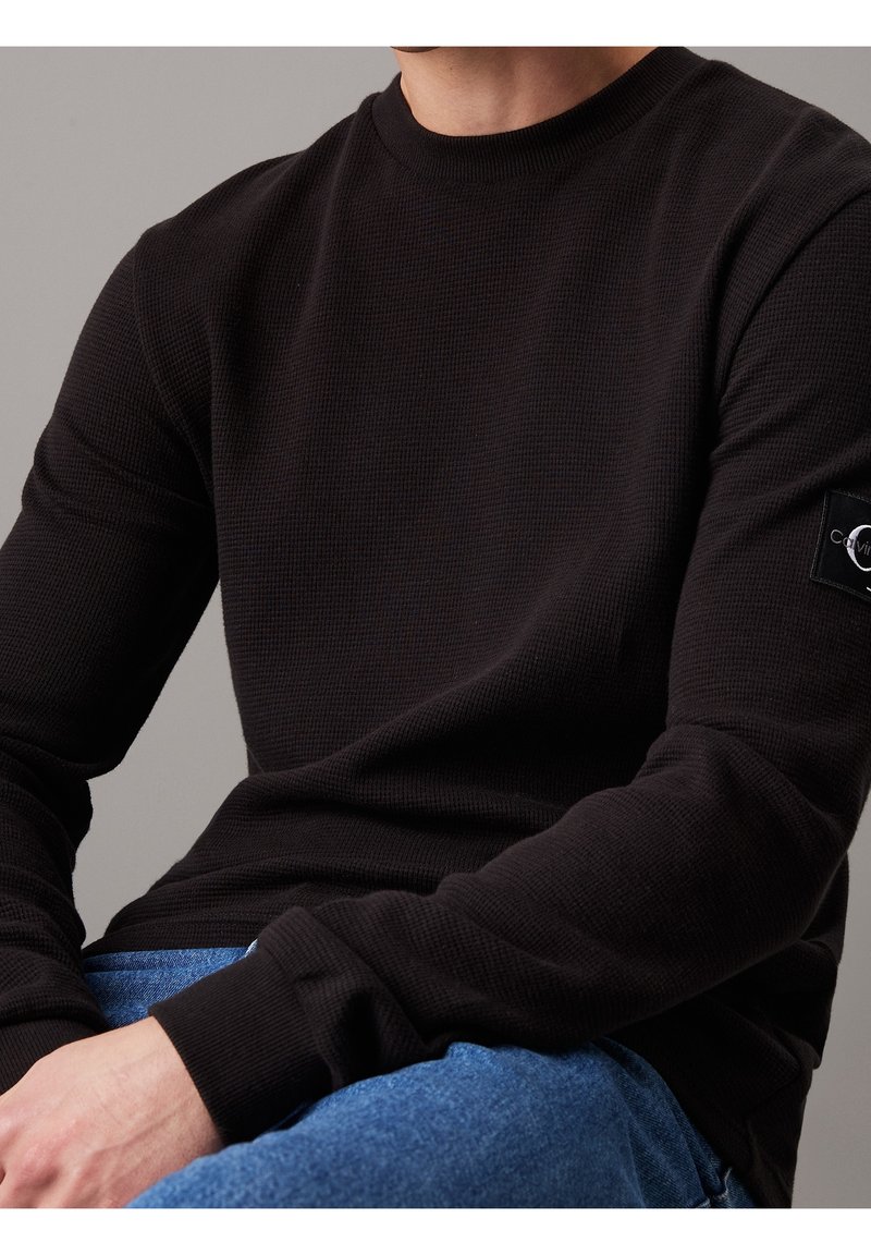 Svart långärmad termoshirt i texturerat tyg, rund hals, och med ett logotyp-märke på ärmen, i kombination med blå denimjeans.