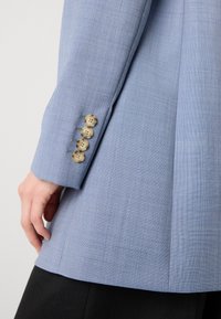 Svetlomodrý blazer s textúrovanou látkou. Obsahuje štyri zlaté gombíky na rukávoch a má prispôsobený strih s vystuženými ramenami.