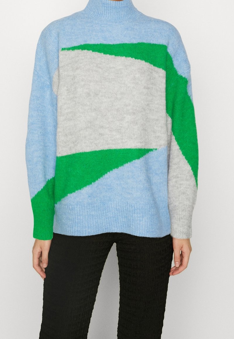 Personne portant un pull à col roulé ample à motif géométrique bleu clair, vert et gris, avec un pantalon noir texturé.