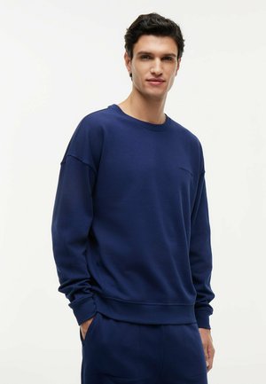 Jeune homme debout les mains dans les poches, portant un sweat-shirt à manches longues bleu marine et un pantalon assorti, sur un fond clair uni.