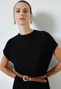 Vestito a maglia nero a coste con maniche corte, strizzato in vita da una cintura in pelle marrone con fibbia in metallo dorato. Orecchini dorati adornano il modello.
