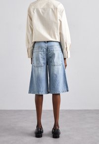 Los culottes de mezclilla azul claro presentan un corte relajado, dos bolsillos traseros y un dobladillo ligeramente acampanado, combinados con una camisa de manga larga color crema.