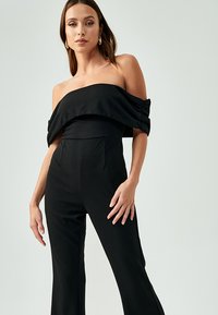 Sort off-the-shoulder jumpsuit med tætsiddende liv, vide ben og sammensunkne ærmer, lavet af et glat stof.