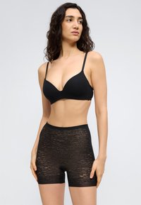 Γυναίκα που φοράει μαύρο σουτιέν με λεπτές τιράντες και δαντελένια σορτς με ψηλή μέση, στέκεται μπροστά σε απλό φωτεινό φόντο, κοιτάζοντας προς τα πλάγια.