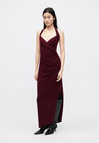 Burgundské maxi šaty s halter výstřihem, s řasenou texturou, hlubokým výstřihem do V a bočním rozparkem, doplněné černými kozačkami sahajícími ke kolenům.