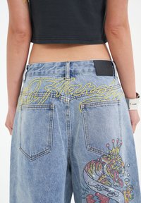 Jeans de mezclilla azul claro con texto bordado amarillo "Ed Hardy" y gráficos coloridos de tigres con brillante en los bolsillos traseros.