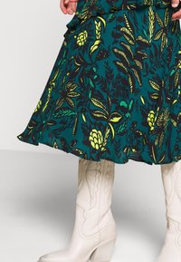 Groene bloemenrok met zwart en gele bladmotieven, met een gerafeld zoom. Gepaard met beige cowboylaarzen met reliëfdetails.
