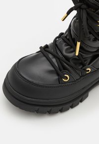 Stivale imbottito nero con punta arrotondata, occhielli in tonalità oro e lacci neri. Presenta una suola in gomma spessa e texture per una migliore aderenza.