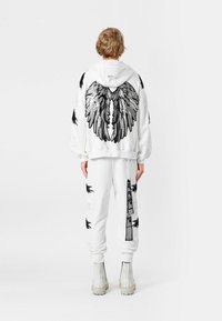 Sudadera blanca con un gráfico de alas negras y detalles desgastados, combinada con pantalones de chándal blancos a juego con acentos y etiquetas, usados con botas.