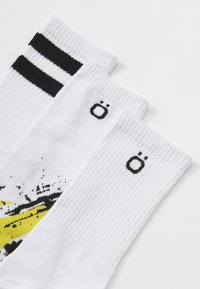 Tres pares de calcetines blancos, con textura acanalada, rayas negras y un diseño gráfico con acentos amarillos y negros. Logotipo mostrado.