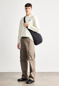 Helle beige Langarmbluse mit blauer Stickerei, kombiniert mit sandfarbenen Cargo-Hosen mit seitlichen Taschen und schwarzen Slippern. Schwarze Umhängetasche.