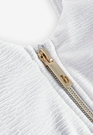 Tessuto bianco testurizzato che mette in mostra una zip dorata con un tirante rettangolare. Il tessuto presenta un delicato motivo ondulato che ne esalta la profondità.