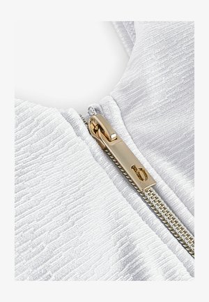 Tissu blanc texturé mettant en valeur une fermeture éclair dorée avec un tirette rectangulaire. Le tissu présente un motif ondulé subtil qui renforce sa profondeur.