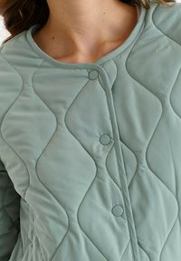 Nahaufnahme einer Person, die eine hellgrüne gesteppte Jacke mit runden Druckknöpfen und einem kragenlosen Ausschnitt trägt.
