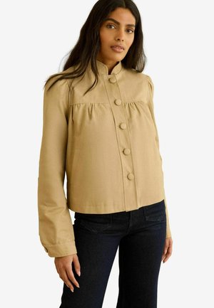 Woman wearing a beige buttoned jacket with puffed shoulders and black pants, standing against a white background.