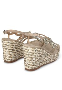 Beżowe sandały na koturnie z plecionym obcasem przypominającym słomę, ozdobne akcenty z cyrkoniami oraz ażurowy design z regulowanym zapięciem na kostce.