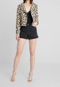 Kurtka motocyklowa z futra w cętki, czarna koszulka na ramiączkach, szorty dżinsowe z wysokim stanem w kolorze czarnym oraz jasne sandały na cienkich obcasach. Proste tło.