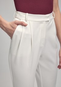 White tailored trousers with front pleats, straight legs, and side pockets. Worn with a ribbed burgundy top, showcasing a smooth texture.