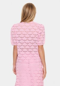 Top in pizzo rosa con maniche corte a sbuffo, caratterizzato da un motivo scallop, texture semitrasparente e scollatura rotonda. Abbinato a una gonna coordinata.