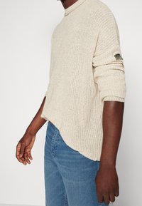 Mann, der einen cremefarbenen gestrickten Pullover mit hochgekrempelten Ärmeln und blaue Jeans trägt, von der Mitte des Oberkörpers bis zur Mitte des Oberschenkels dargestellt.