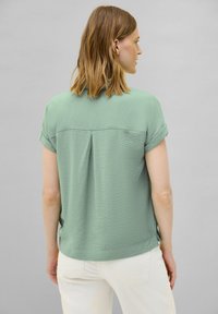 Blusa a maniche corte di colore verde chiaro in tessuto testurizzato, con cucitura a yoke sul retro e maniche arrotolate, abbinata a jeans bianchi.