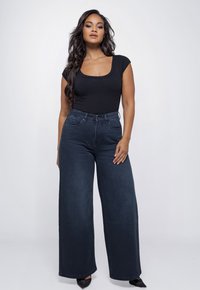 Vrouw met lang golvend haar, gekleed in een zwarte top met ronde hals, hooggerimpelde, wijde donkere spijkerbroek en zwarte puntige schoenen.