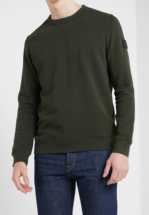 Mann trägt dunkelgrünes Sweatshirt mit Rundhalsausschnitt und langen Ärmeln sowie blaue Jeans, stehend vor neutralem Hintergrund.