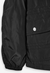 Czarna kurtka puchowa o gładkim, błyszczącym wykończeniu, z wyraźnymi szwami i zapięciem na zatrzask przy kieszeni na piersi. Elastyczne mankiety.