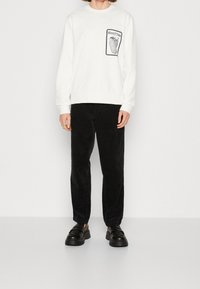 Vit sweatshirt med grafisk framficka, svarta manchesterbyxor och svarta platåskor, uppvisat mot en neutral bakgrund.
