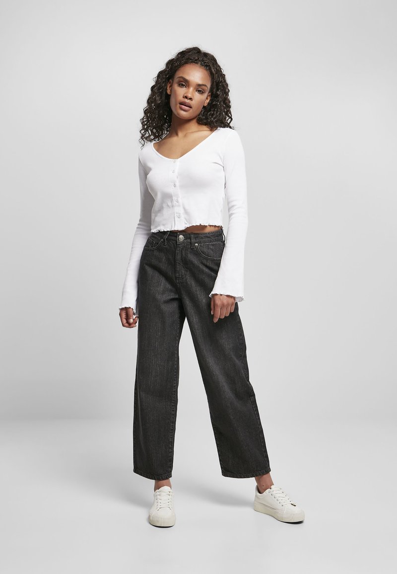 Witte lange mouwen crop top met knoopsluiting en rafelige zoom, gecombineerd met zwarte wijde jeans en witte sneakers. Neutrale achtergrond.