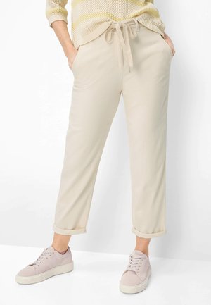 Persona che indossa pantaloni beige con coulisse arrotolati alle caviglie, con le mani nelle tasche e sneaker rosa chiaro su sfondo bianco.