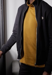 Schwarze Zip-Jacke mit hohem Kragen und strukturierten Ärmeln, über einem senfgelben Rollkragenpullover getragen. Die Jacke hat ein detailliertes Emblem.