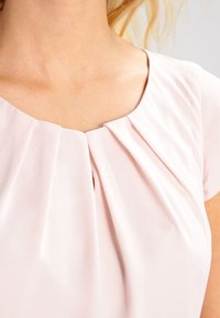Kremsrosa blus med korta ärmar, med en plissérdesign vid halsringningen och ett lent, lättviktigt tyg.