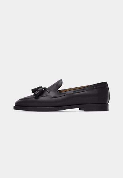 Schwarzer Herren-Loafer aus Leder mit Quasten oben, niedrigem Absatz und dekorativer Naht entlang der Seiten vor einfarbigem Hintergrund.