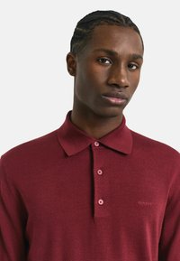 Polo shirt bordeaux en tissu léger ; dispose d'un col, de trois boutons et d'une broderie de logo ton sur ton sur la poitrine.