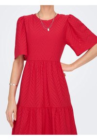 Femme portant une robe rouge à œillets avec des manches bouffantes, un collier en argent et un bracelet en argent, debout avec une main sur la hanche.