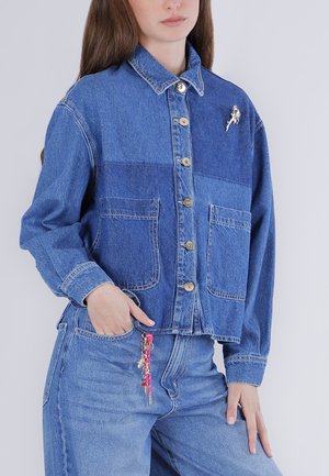 Vrouw draagt een blauw patchwork spijkerjasje met grote voorzakken en een zilveren astronautenbroche, blauwe spijkerbroek met een rood sleutelhangeraccessoire.