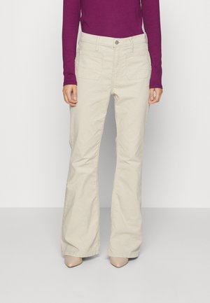 Person trägt beige ausgestellte Hose, lila Langarmshirt und beigen spitzen Schuhe, steht vor einem schlichten weißen Hintergrund.