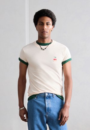 Młody mężczyzna z krótkimi kręconymi włosami, w kremowym T-shircie z podwiniętymi zielonymi rękawami, z logo wiśni, nałożonym na zieloną koszulę i niebieskie dżinsy.