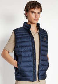 Gilet imbottito blu navy con colletto alto, trapuntatura orizzontale, chiusura con zip e tasche laterali; indossato sopra una camicia beige a maniche corte.