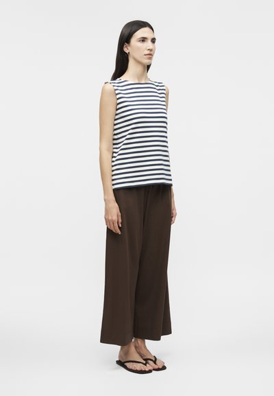 Γυναίκα που στέκεται φορώντας ένα αμάνικο μπλε-σα navy με λευκές ρίγες τοπ, φαρδιά σκούρα καφέ παντελόνια με φαρδύ πόδι και μαύρα σαγιονάρες σε λευκό φόντο.