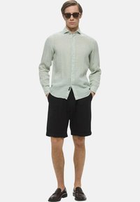 Camicia button-up in lino verde chiaro e pantaloni corti neri arrotolati. Il modello indossa anche occhiali da sole scuri e mocassini scuri. Design semplice e vestibilità rilassata.
