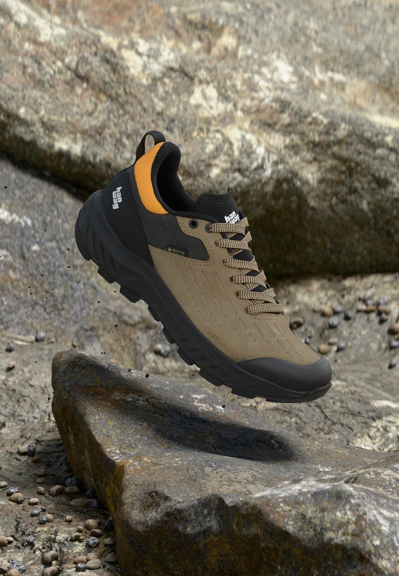 Hněro-černá turistická bota s visačkou Gore-Tex, oranžovým detailem na patě a robustní podrážkou, zavěšená nad kamenitým terénem.