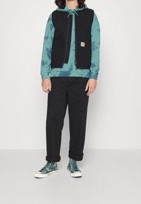 Černá bezrukávová vesta přes kapucí s teal tie-dye vzorem, spárovaná s černými kalhotami a vzorovanými zelenými vysokými teniskami. Viditelné zapínání na zip a kapsy.